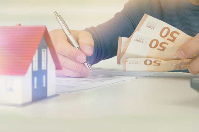 Geldscheine und ein Stift in der Hand. Nebenan ist ein kleines Modellhaus abgebildet